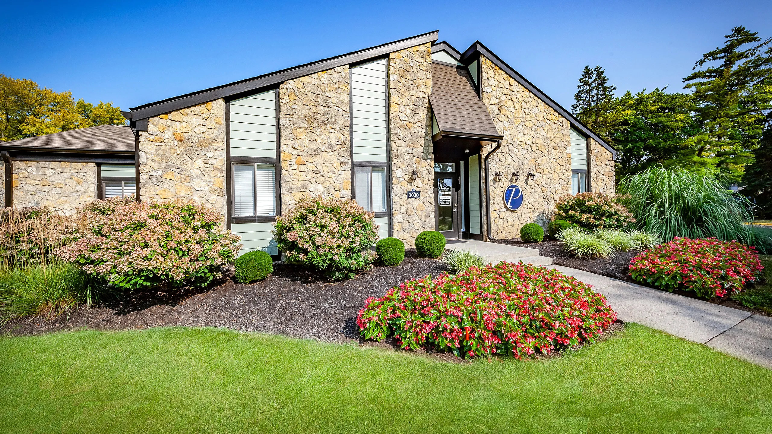 Charming stone-clad leasing office with beautiful landscaping and vibrant flower beds under a clear blue sky.