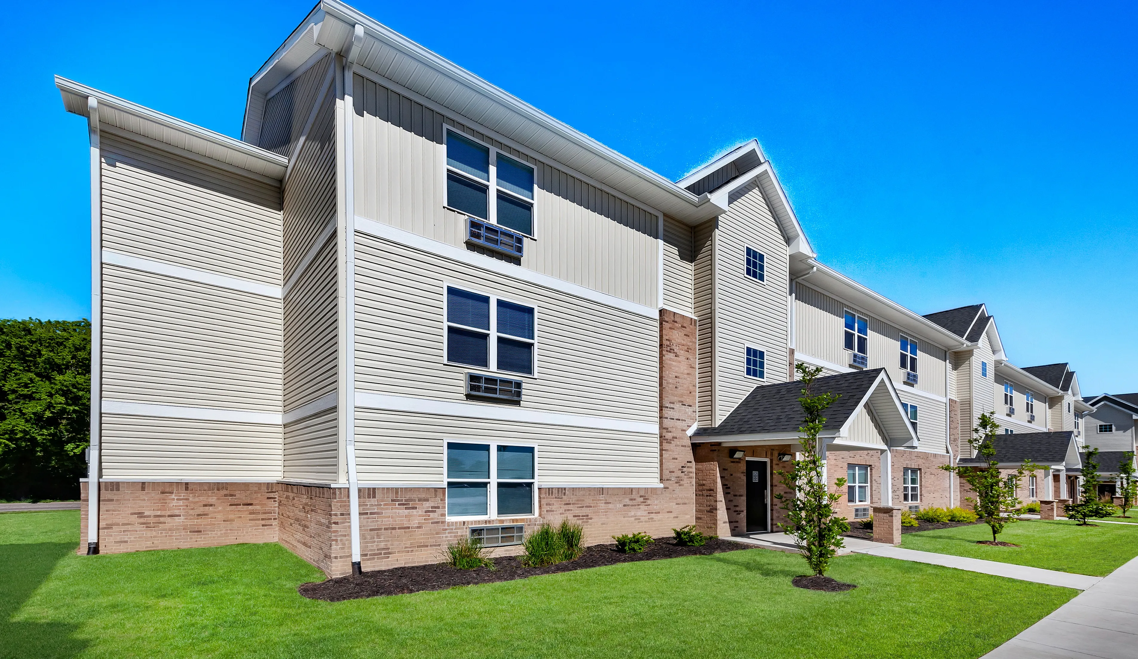 Modern apartment complex surrounded by lush green lawns and clear blue skies, featuring contemporary architecture with brick and siding design.