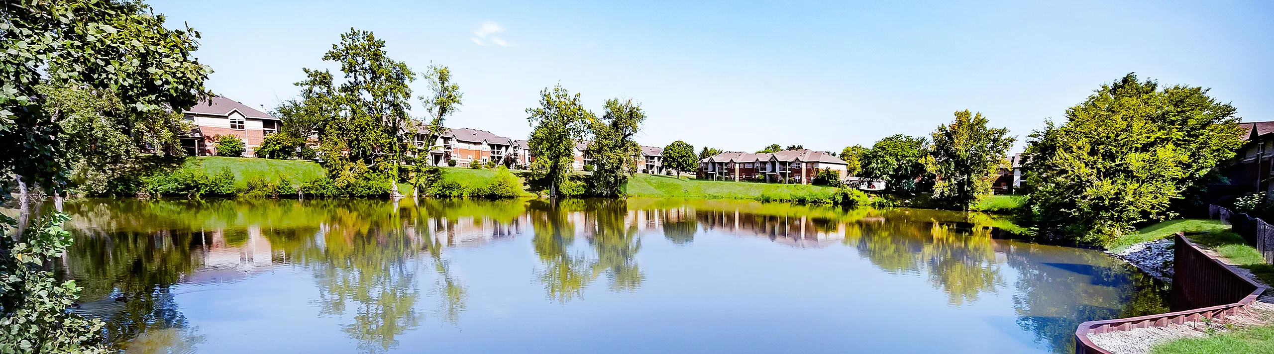 Charming lakeside apartment view showcasing lush greenery and tranquil water reflections under a bright blue sky.