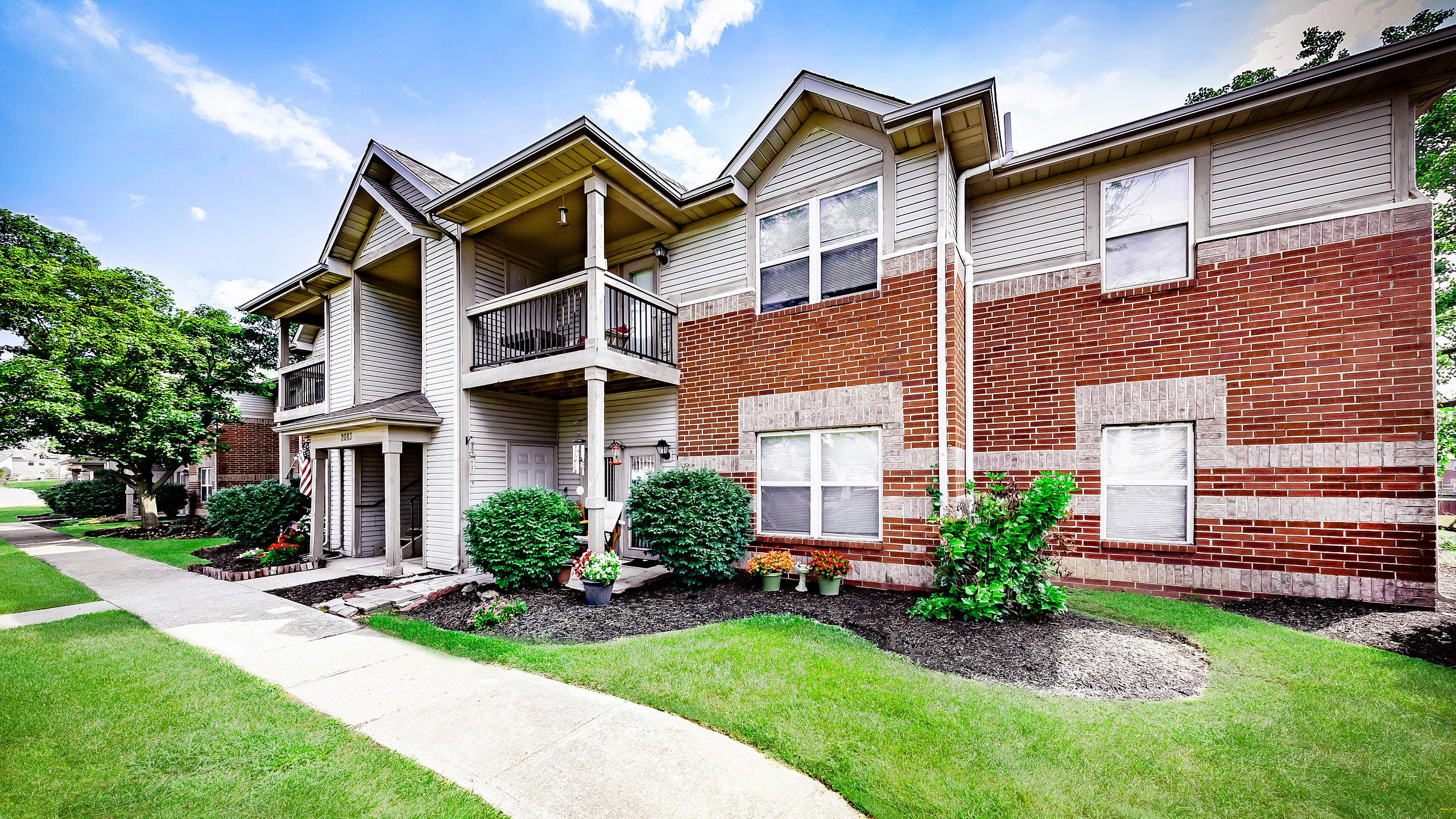 Greenfield Crossing Apartments - Charming two-story apartment building with red brick and beige siding, surrounded by lush greenery and a bright blue sky.