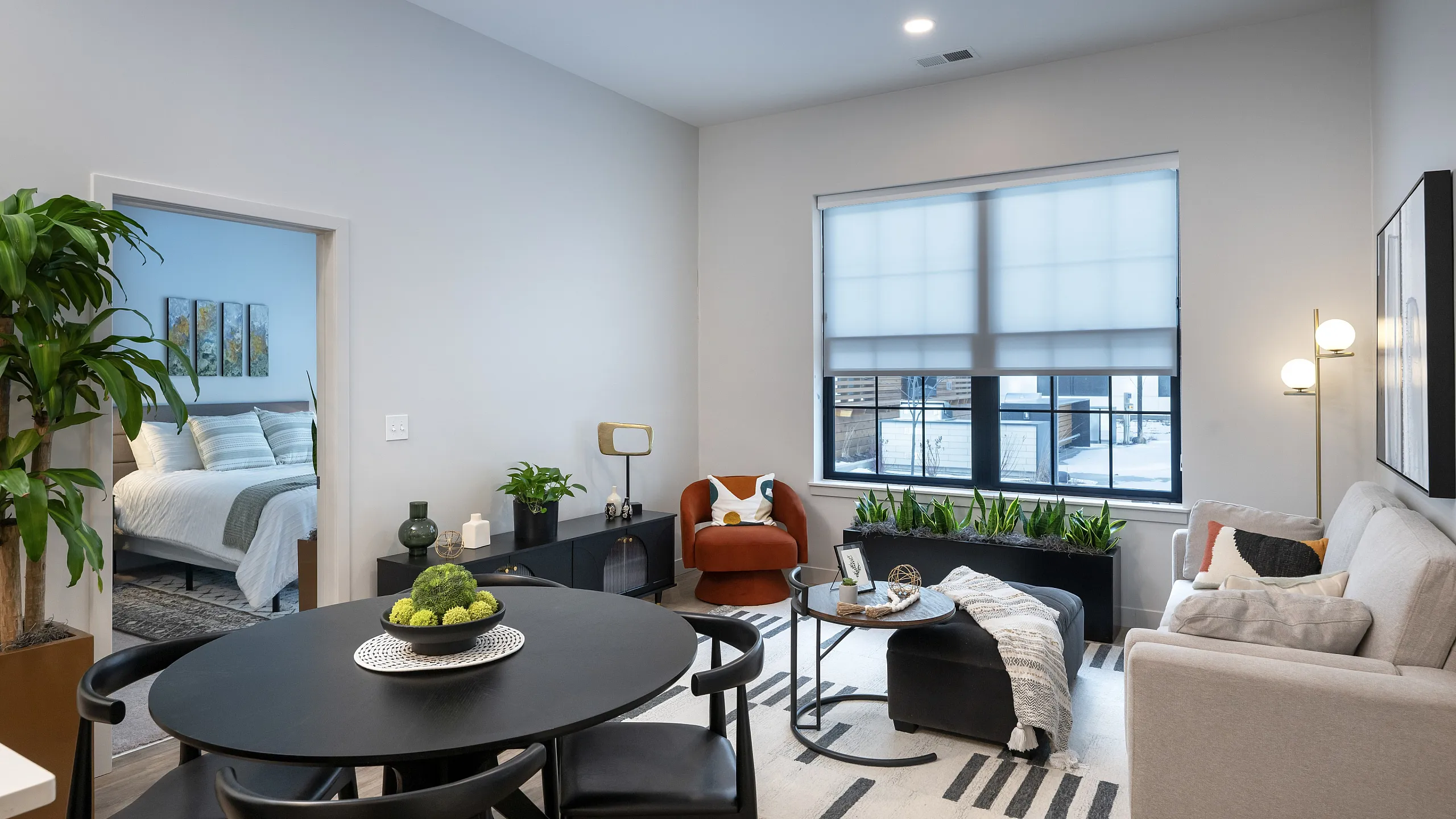 Modern living room with stylish decor, soft gray sofa, vibrant orange chair, and lush greenery, adjacent to a cozy bedroom entrance.