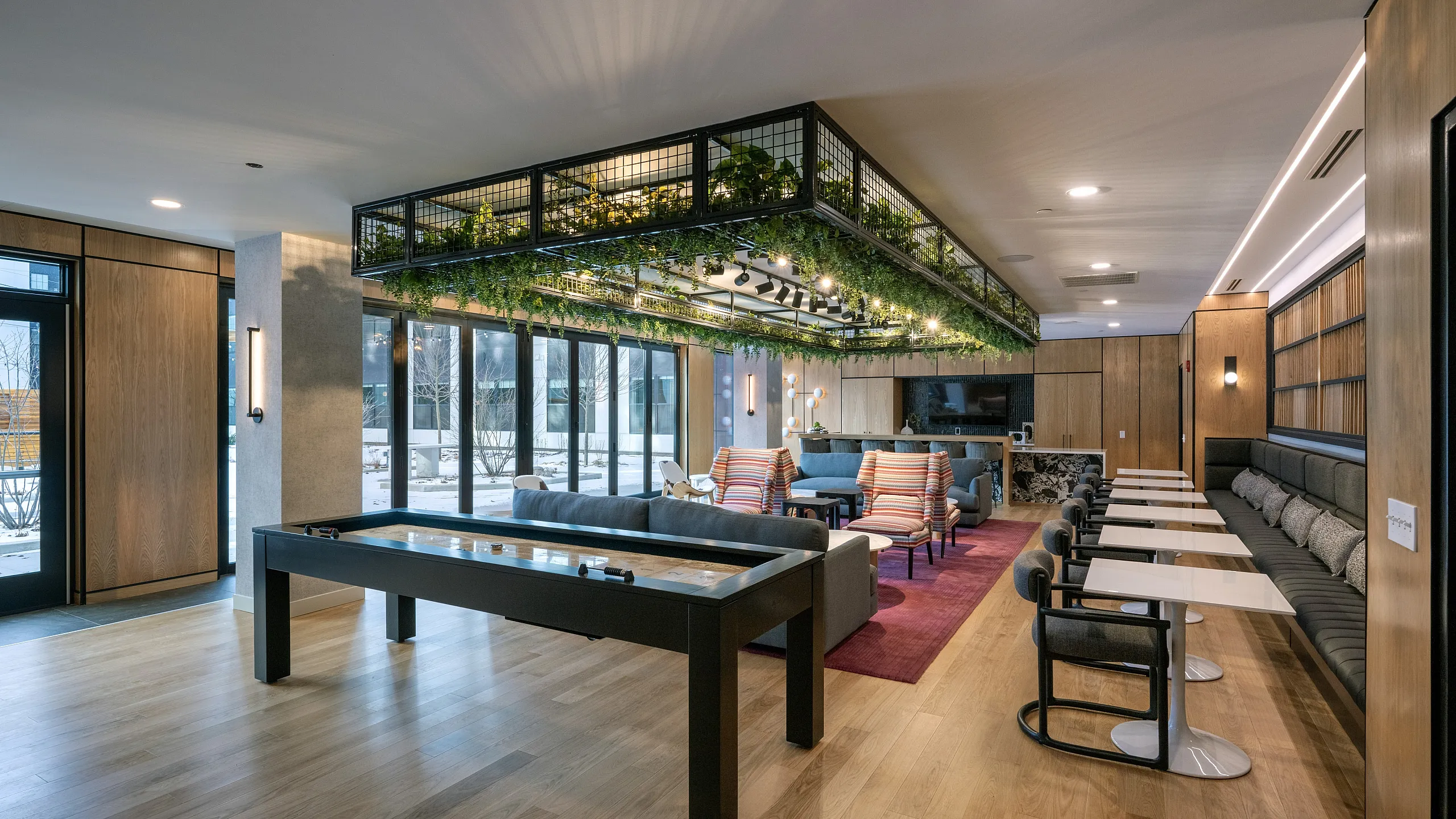 The Elex: Modern communal lounge featuring sleek wooden floors, cozy seating areas, vibrant greenery, and a game table.