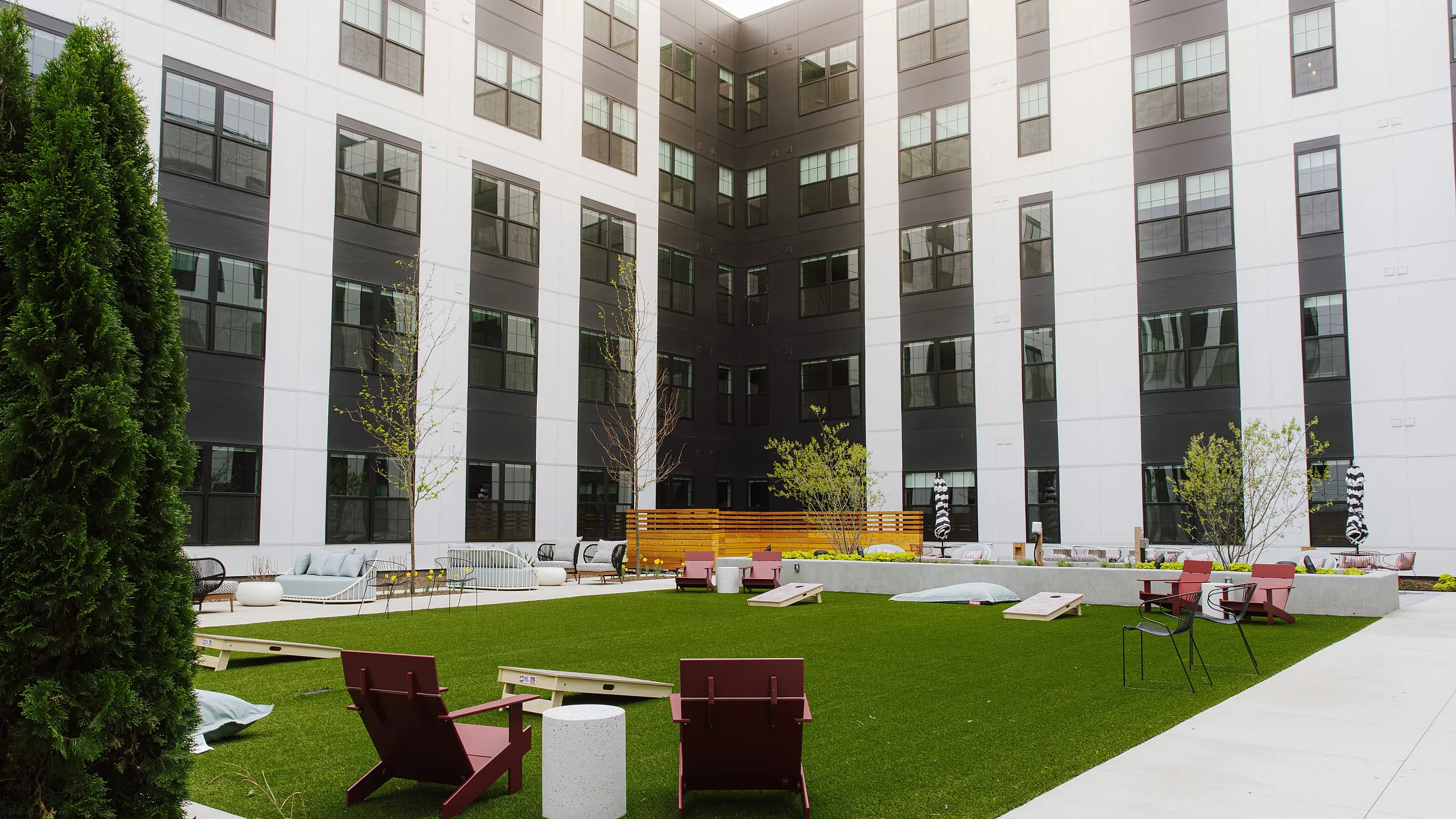 Vibrant apartment courtyard featuring lush green lawns, stylish seating, and modern outdoor games, nestled between sleek, multi-story buildings.