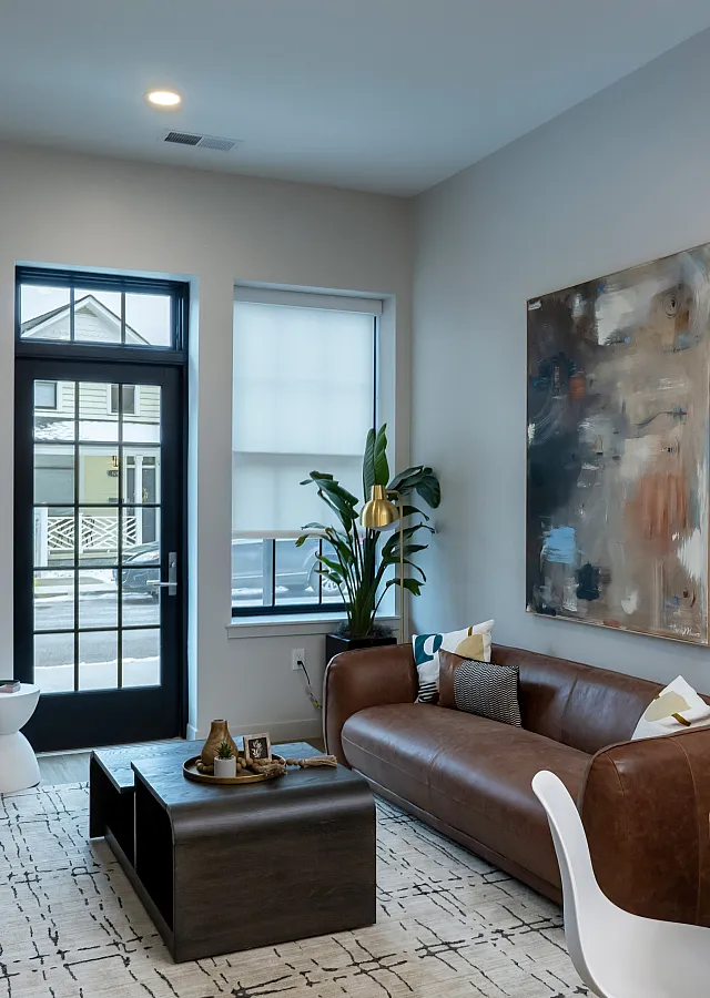 The ELex: Modern living room with elegant decor, featuring a cozy brown leather sofa, abstract artwork, and abundant natural light through large windows.