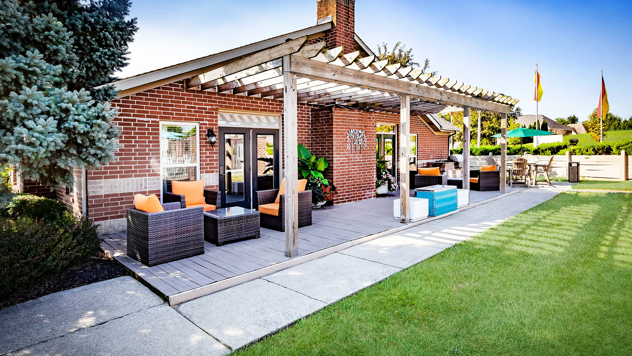 Inviting outdoor patio with stylish wicker furniture and vibrant orange cushions, set against a charming brick clubhouse under a wooden pergola.