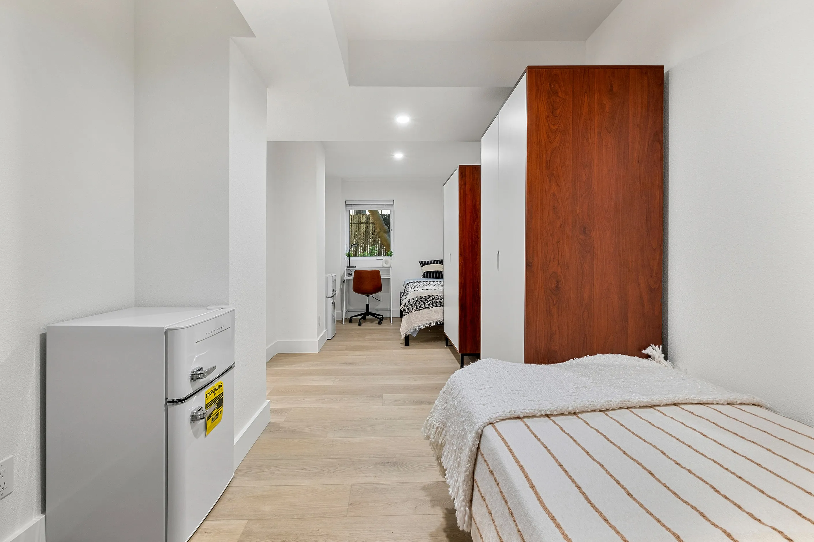 Bright bedroom featuring a modern design with sleek wooden wardrobes, cozy bed, and a compact white refrigerator, ideal for comfortable living.