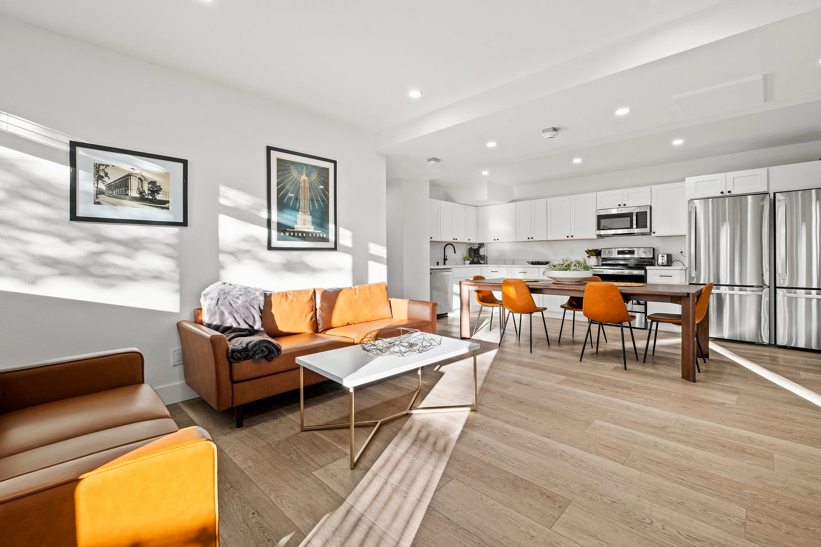 Bright and modern open-plan living room with stylish leather sofas, a sleek coffee table, and an inviting dining area. The contemporary kitchen features stainless steel appliances and ample storage, highlighted by natural wood flooring and artistic decor.