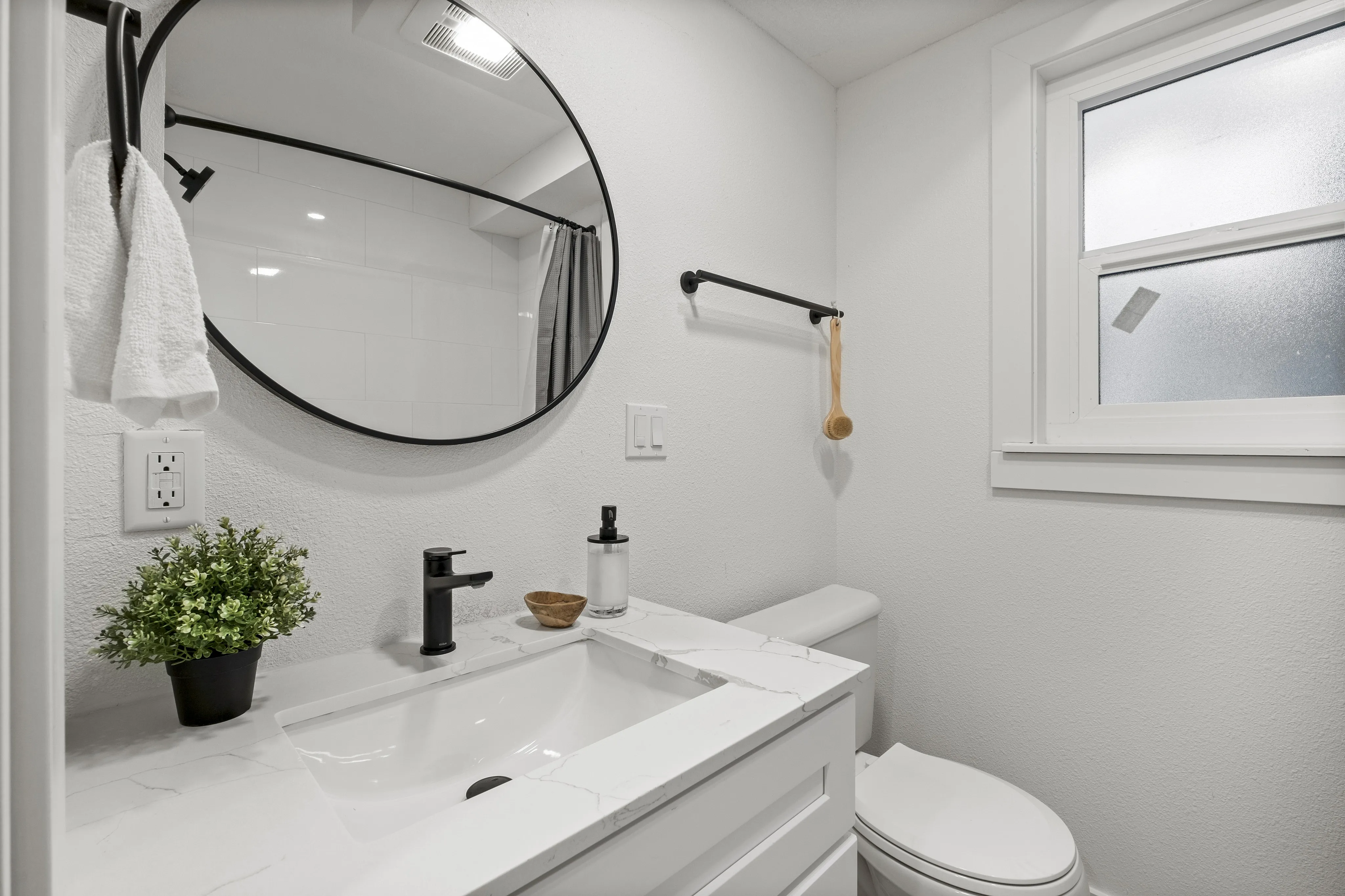 Elegant modern bathroom with a chic round mirror, sleek black fixtures, and a bright window illuminating a stylish vanity.