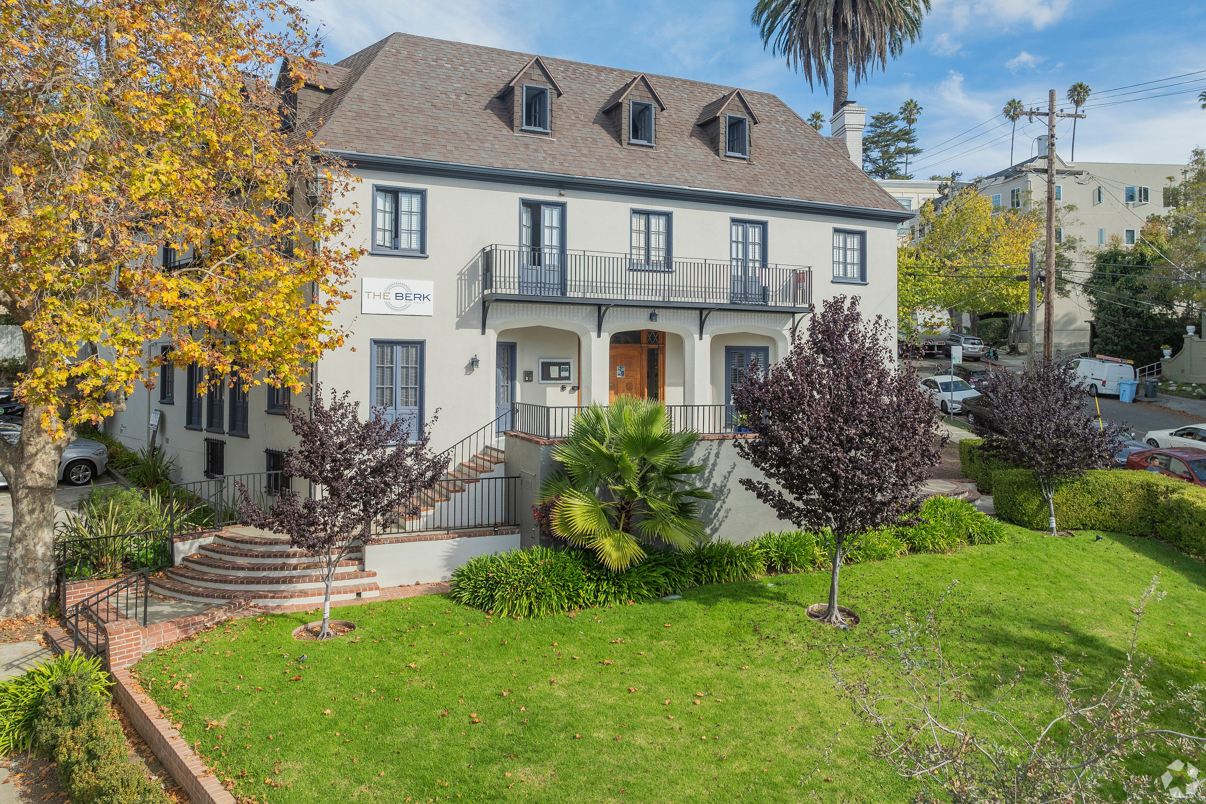 Charming three-story residential building with elegant dormer windows, lush greenery, and a welcoming staircase entrance.