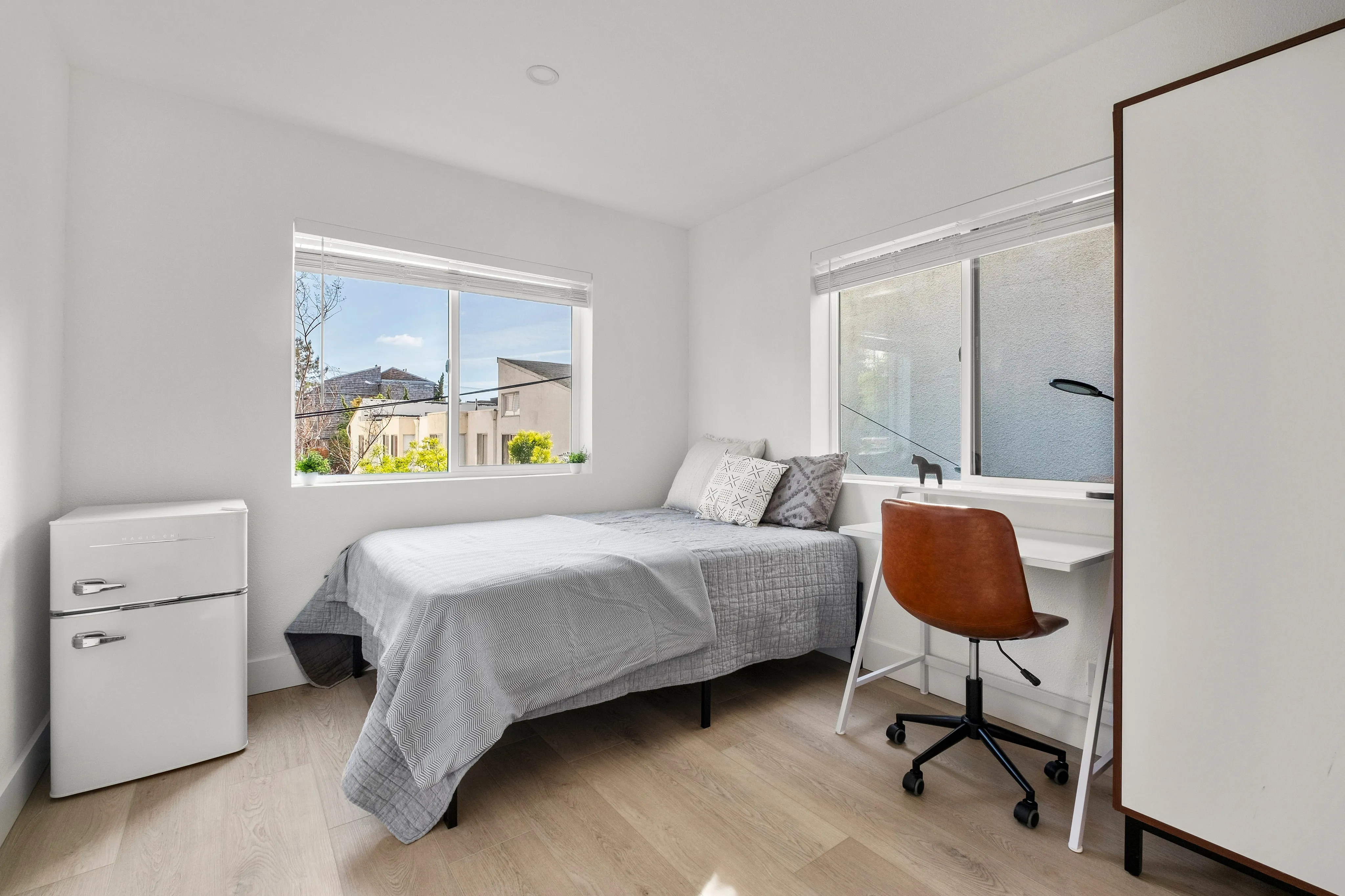 Bright and inviting bedroom with a cozy bed, modern desk, and stylish chair, enhanced by large windows offering scenic views.