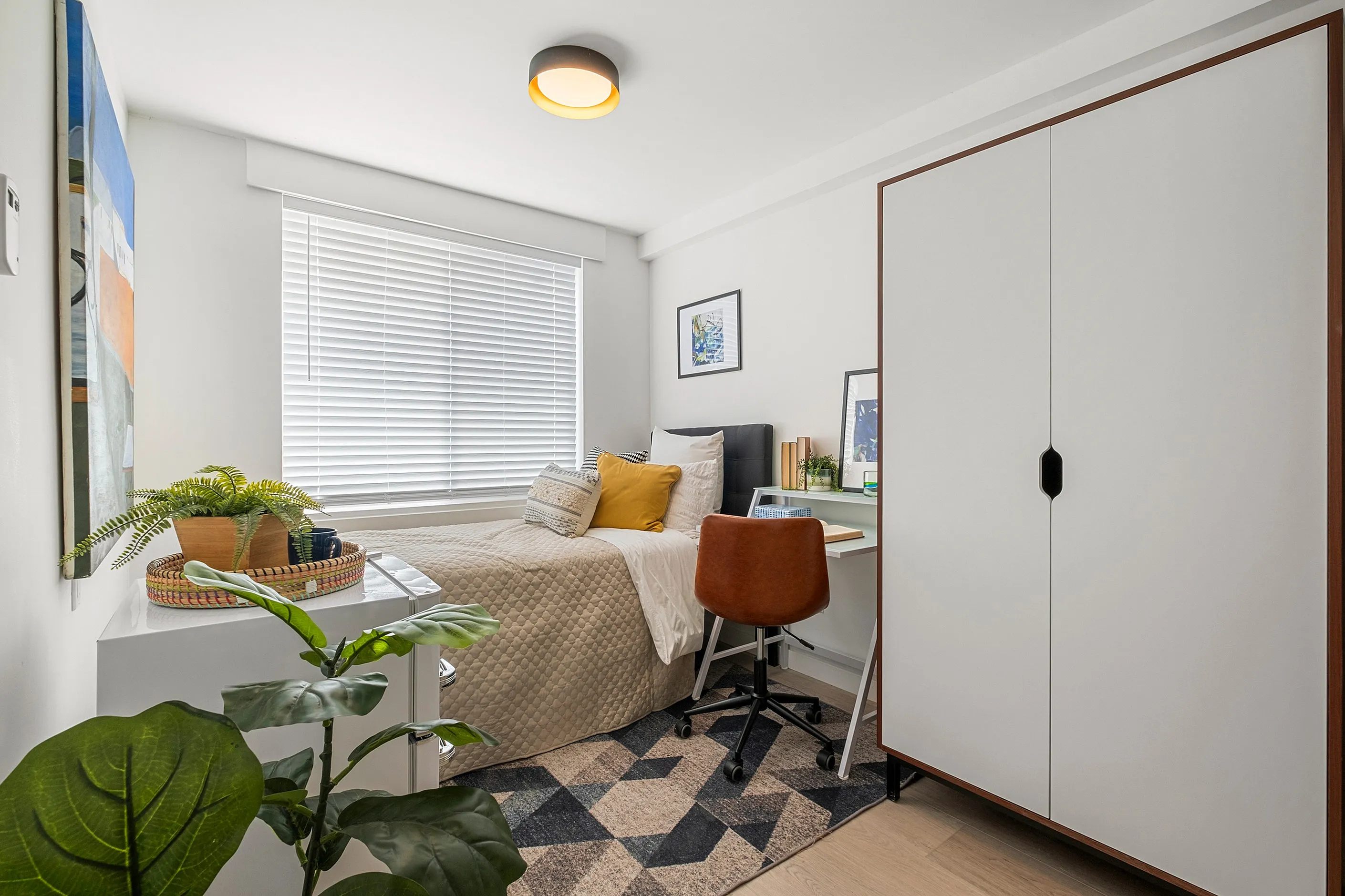 Fully furnished modern bedroom featuring a cozy bed with accent pillows, compact desk with a stylish chair, large wardrobe, and lush houseplant under a soft ceiling light.