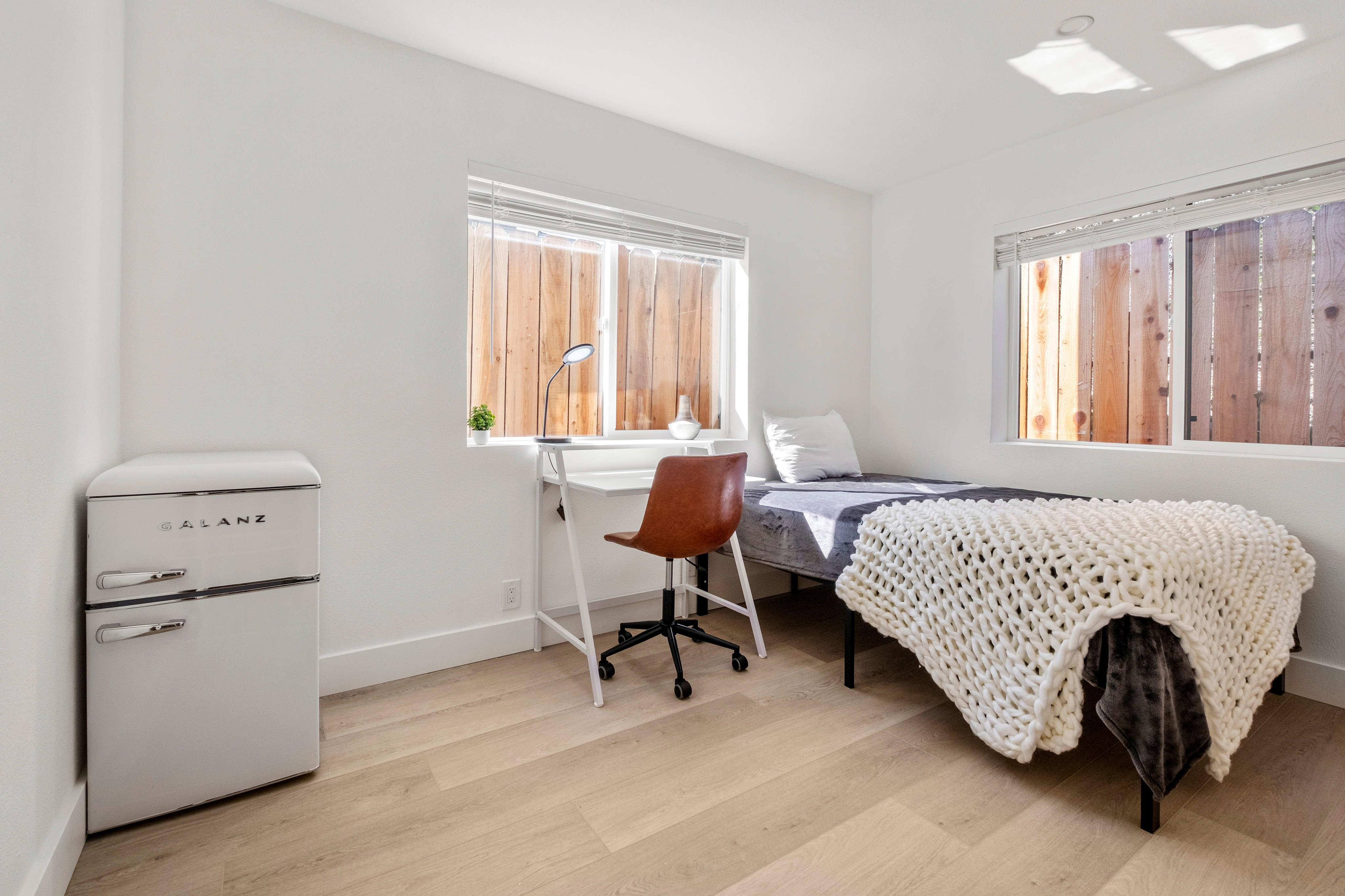 A fully furnished bright and cozy bedroom with modern decor, featuring a compact white fridge, sleek desk, and stylish knitted blanket.