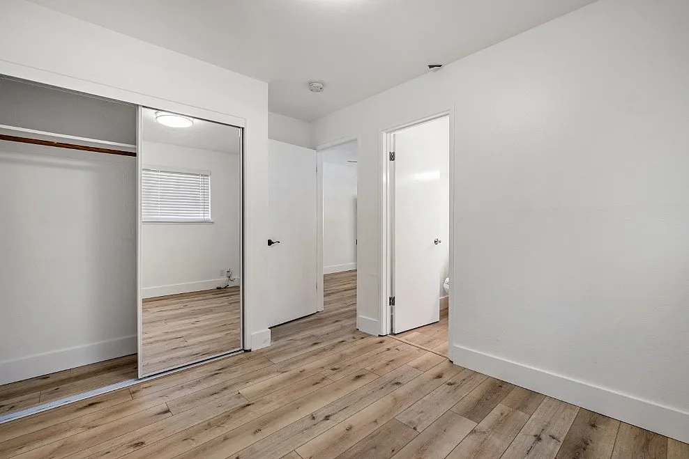 Bright and airy bedroom with sleek wooden flooring, mirrored closet doors, and modern white walls, perfect for stylish living.