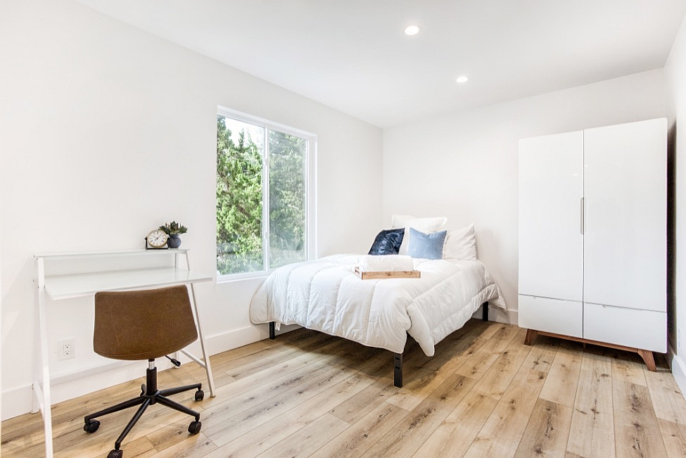 Bright and modern bedroom with large window, featuring cozy white bedding, a sleek desk with a chair, and a stylish wardrobe on elegant wood flooring.