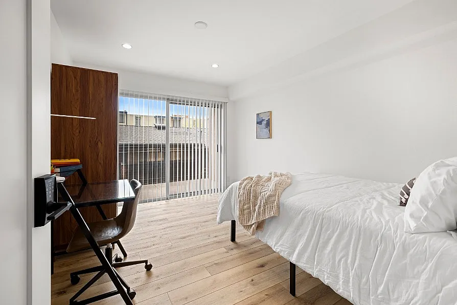 Bright and airy bedroom with modern desk, sleek wooden floor, and large sliding glass doors leading to a serene outdoor view.