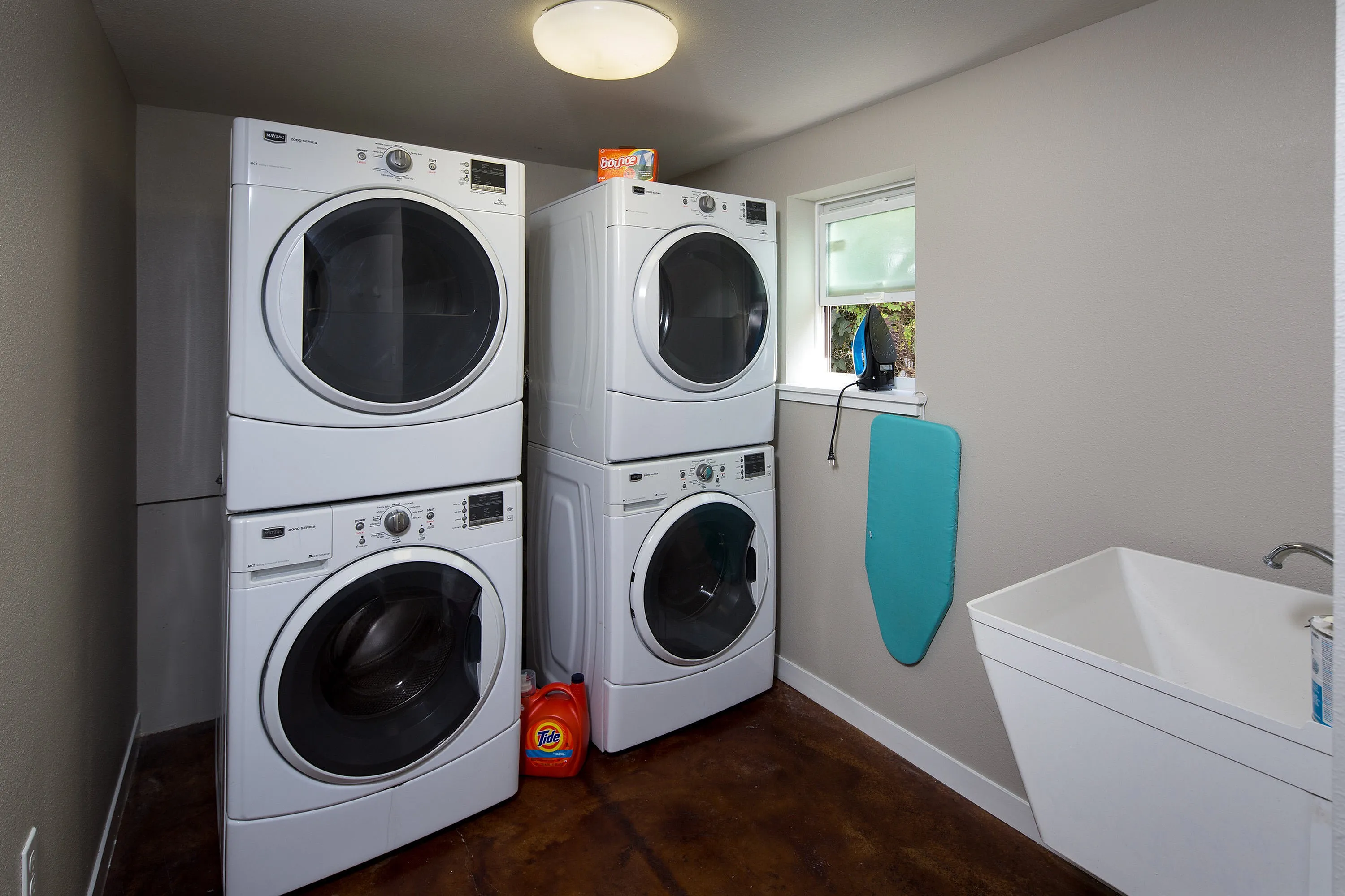 Laundry room featuring dual stackable washers and dryers, a sleek utility sink, and a bright window for natural light.