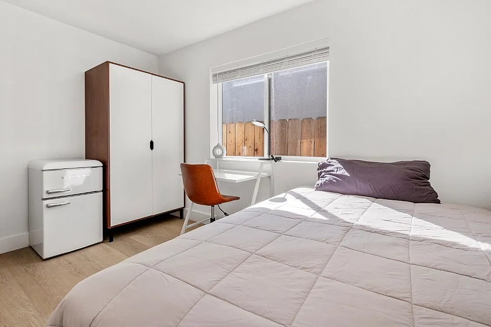Bright and cozy bedroom with contemporary decor, featuring a comfortable bed, sleek wardrobe, and a stylish mini-fridge under natural light.