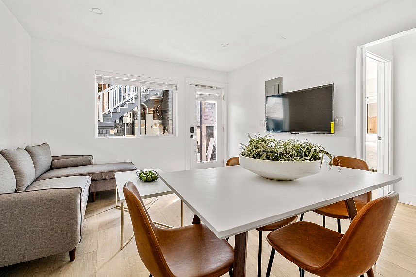 Inviting living space with a modern dining area, sleek leather chairs, and a cozy gray sectional sofa. Sunlit ambiance with a large TV and stylish decor.
