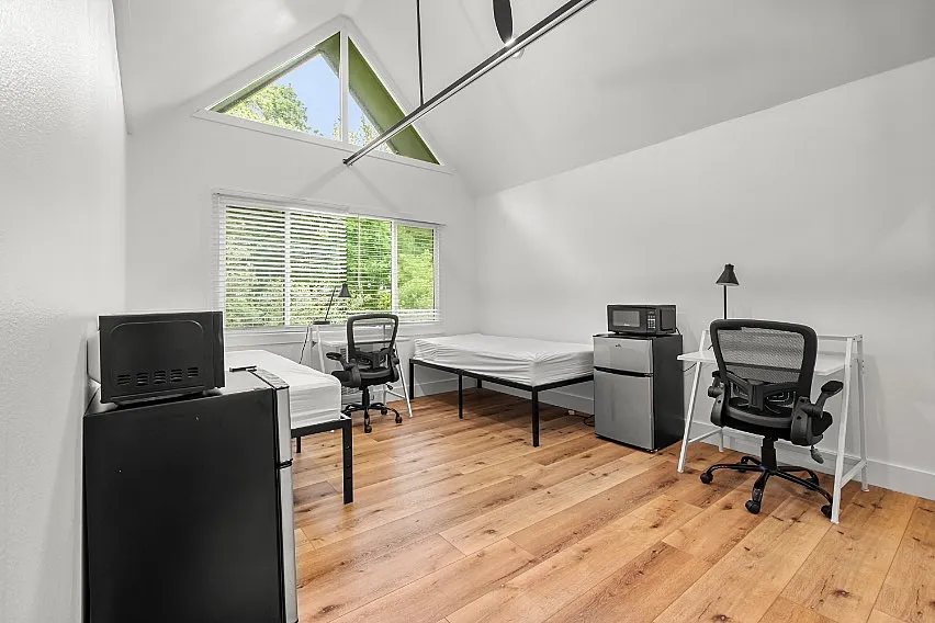 Bright and airy dorm-style room with high ceilings, featuring two single beds, cozy workstations, and modern appliances against a backdrop of lush greenery visible through large windows.