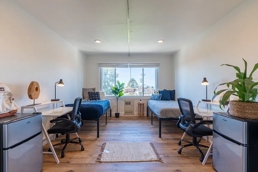 Bright, modern bedroom with two beds, stylish workspaces, and a large window overlooking greenery. Ideal for roommates or a spacious home office.