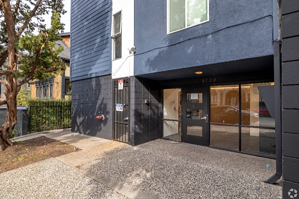 Modern apartment entrance with sleek dark blue facade, large windows, and secure entryway surrounded by greenery.