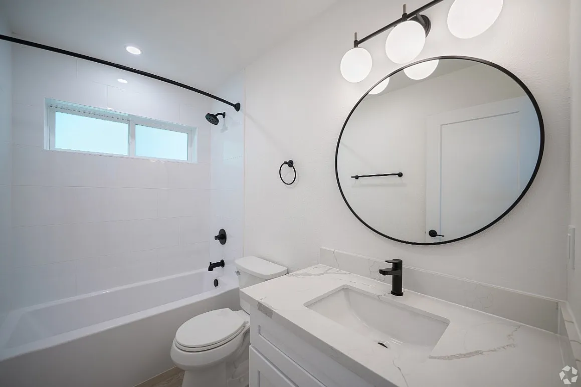 Modern bathroom featuring a sleek white vanity, large round mirror, black fixtures, and a spacious bathtub beneath a bright window.