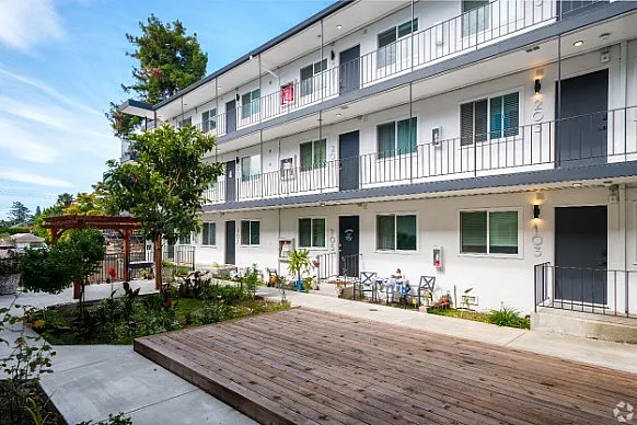 Charming apartment complex exterior featuring three levels of modern living spaces with balconies, lush landscaping, and a cozy courtyard area.