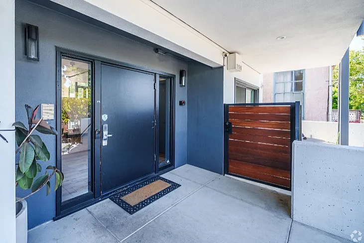 Modern apartment entrance featuring sleek black doors, elegant wood accents, and a welcoming doormat, set within a bright and airy outdoor space.