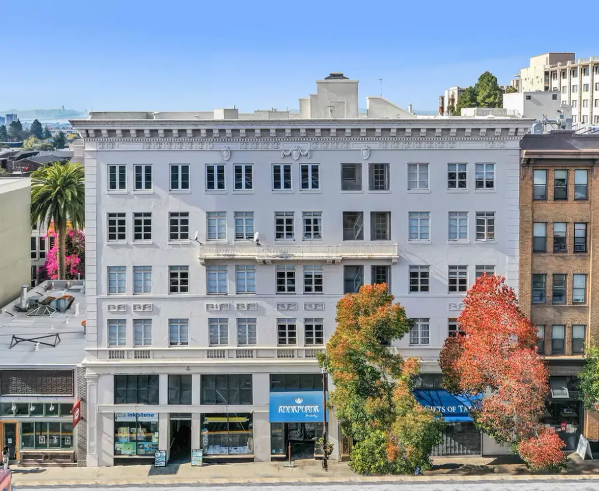 Elegant urban apartment building with classic architecture, vibrant trees, and commercial shops at street level, set against a clear blue sky.