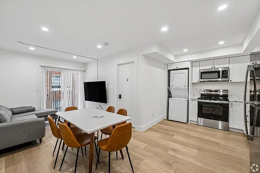 Modern apartment interior featuring an open-plan living area with a sleek kitchen, cozy gray sofa, stylish dining table with brown chairs, and wall-mounted TV. Bright natural light floods through large sliding doors, enhancing the fresh and inviting atmosphere.