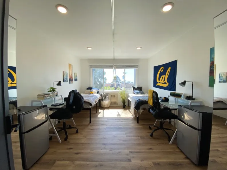 Bright student dorm room featuring two twin beds, large window with a scenic view, wooden flooring, study desks with lamps, and a cozy atmosphere.