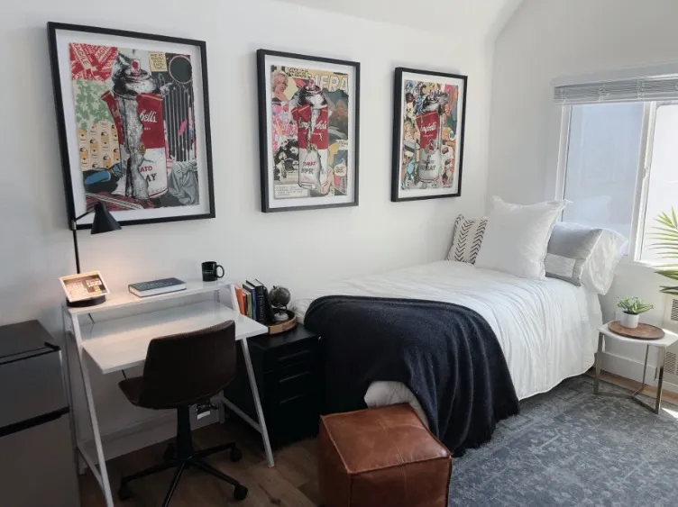 Cozy bedroom with a chic design featuring a single bed, modern artwork on the walls, a sleek white desk with a lamp, and a large window inviting natural light.