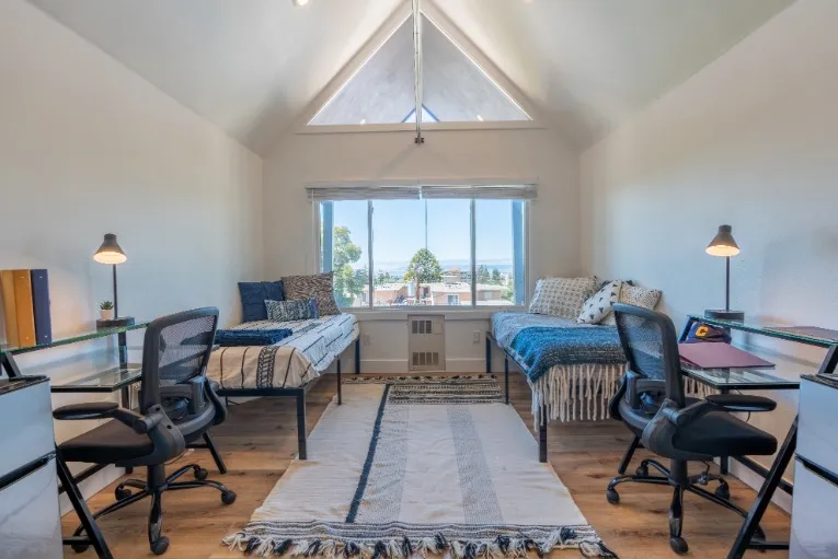 Bright and inviting attic bedroom featuring two cozy beds, modern desks, and a large window with a scenic view.