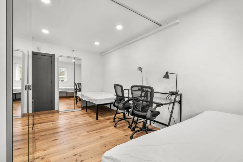 Modern minimalist bedroom with sleek wood flooring, mirrored closet doors, and a functional workspace setup.