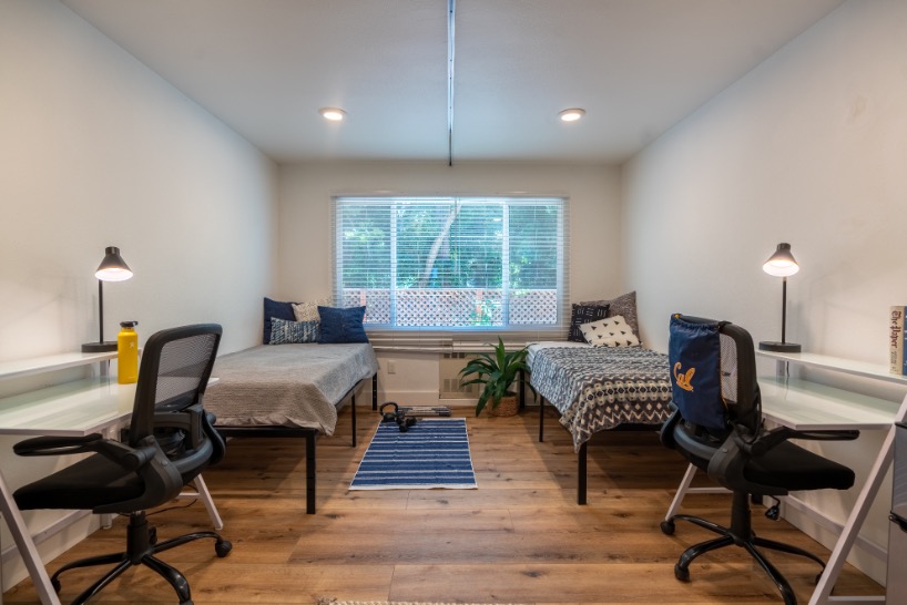 Bright and inviting shared bedroom with two beds, study desks, and a large window overlooking lush greenery, perfect for student living or cohabitation.