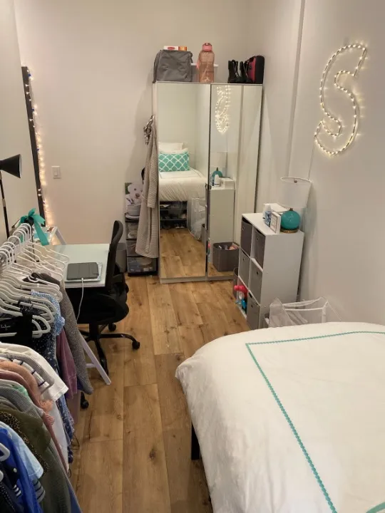 Cozy bedroom with a stylish mirrored wardrobe, inviting bed, elegant string lights, and a functional workspace on warm wood flooring.