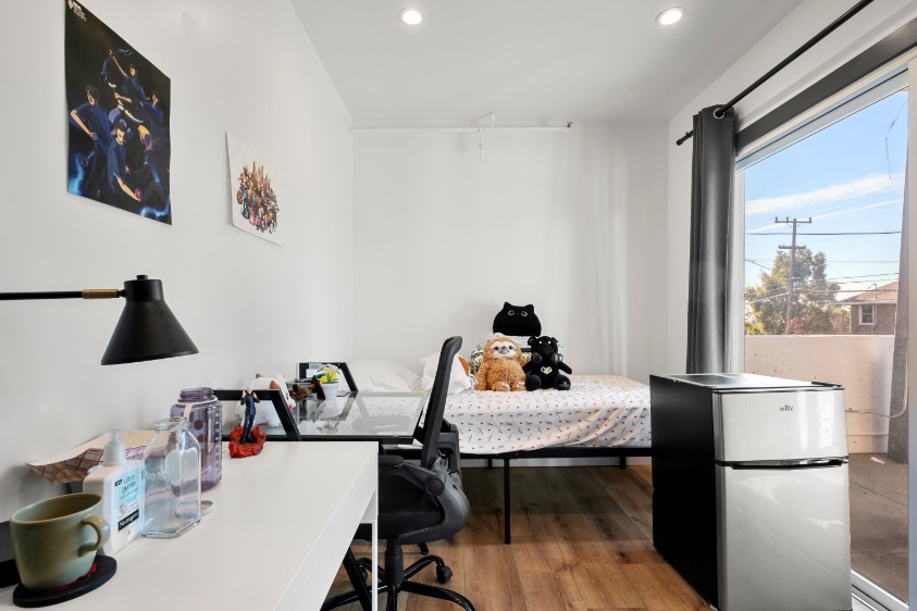 Bright bedroom with stylish decor, featuring plush toys on the bed, a modern desk setup, and a mini fridge by a large window offering ample natural light.