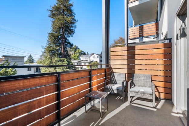 Charming balcony with cozy seating, enhanced by sleek wooden railing and scenic views of lush greenery and nearby buildings.