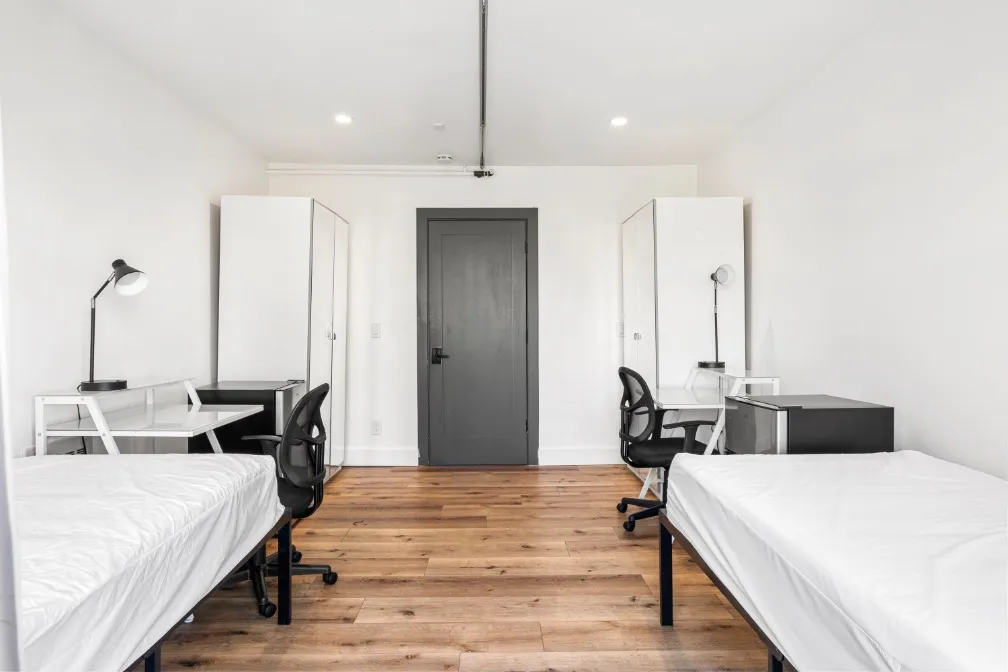Bright and spacious bedroom featuring two comfortable beds, stylish wood flooring, modern desks with chairs, and a contemporary design.