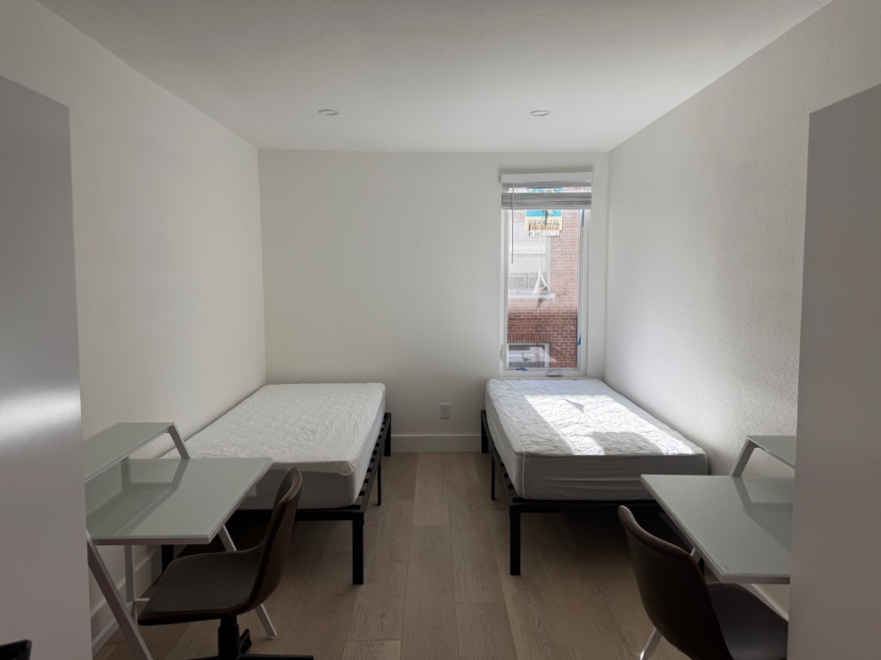 Bright and modern bedroom with two single beds, sleek work desks, and ample natural light from a large window.