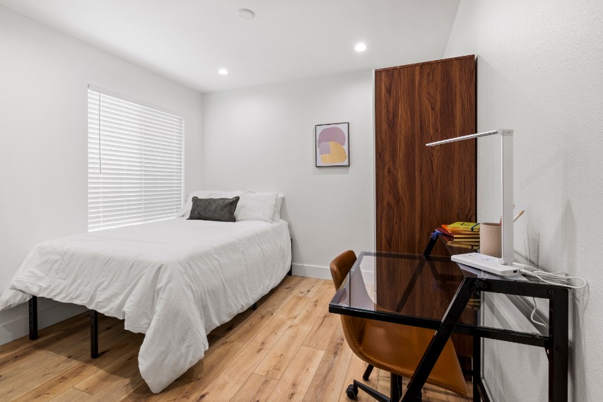 Cozy bedroom with a comfortable bed, sleek desk setup, and stylish wooden wardrobe, featuring warm lighting and modern decor.