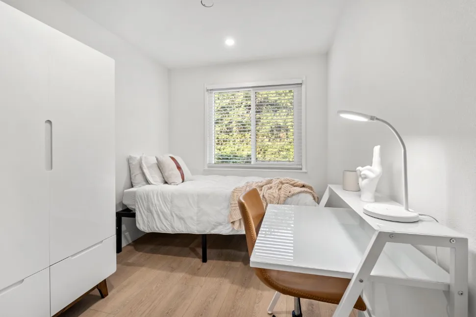 Bright and cozy bedroom with modern furnishings, featuring a comfortable bed, sleek white desk, and natural light from a large window.