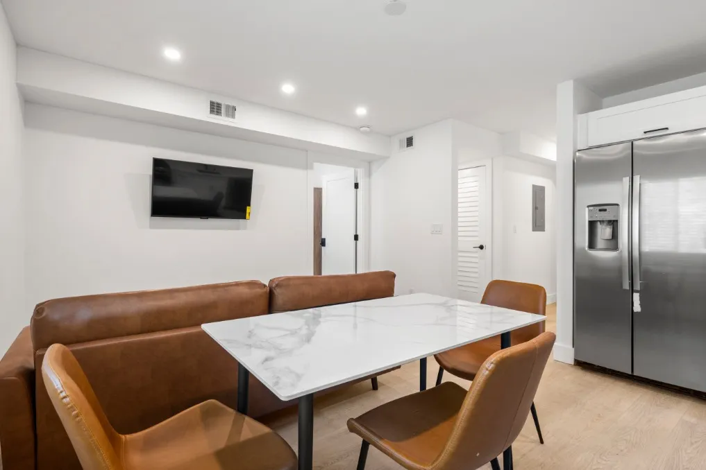 Modern dining area with sleek marble table, brown leather seating, wall-mounted TV, and stainless steel fridge.