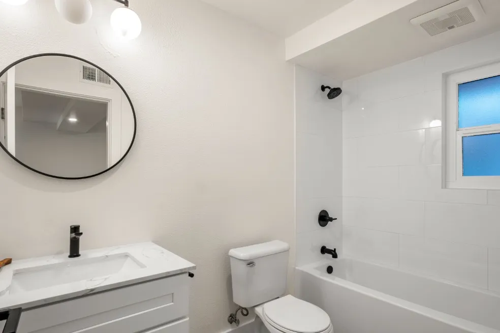 Sleek and modern bathroom featuring a white vanity with a marble countertop, round mirror, elegant fixtures, and a bright shower area with a window.