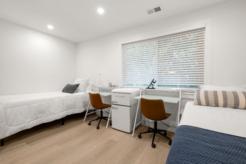 Bright and modern bedroom with two cozy single beds, sleek desks with chairs, and a compact mini fridge, perfect for a comfortable and practical living space.