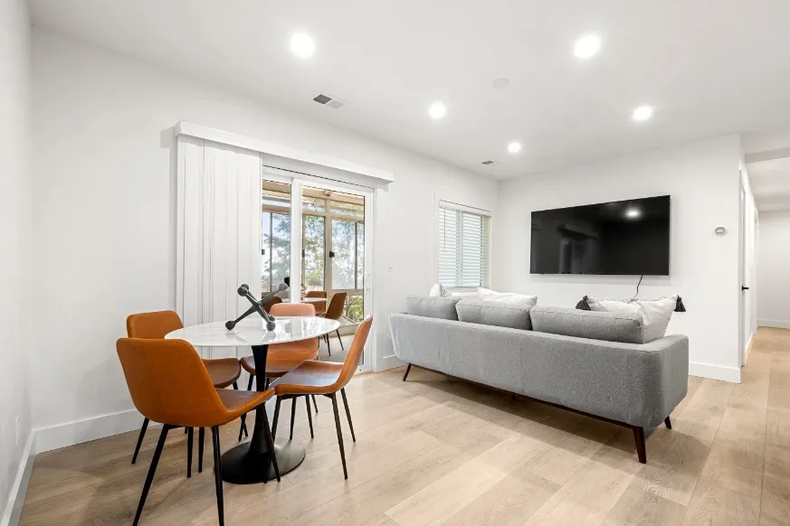 Bright and modern living area featuring a sleek gray sofa, wall-mounted TV, and stylish dining set with orange chairs, accented by natural light from large windows.