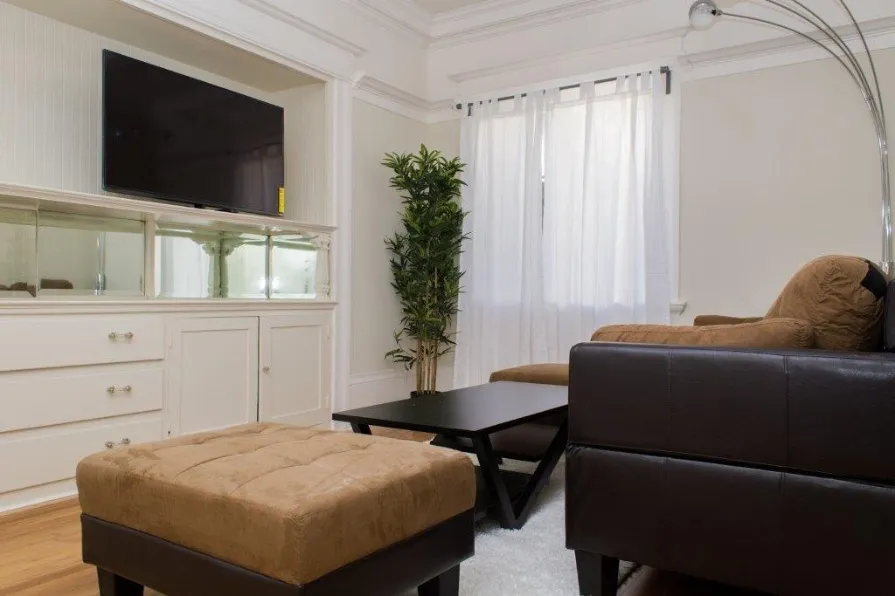 Elegant living room featuring a sleek entertainment center with wall-mounted TV, plush brown seating, and a stylish floor lamp, complemented by natural light through sheer curtains.