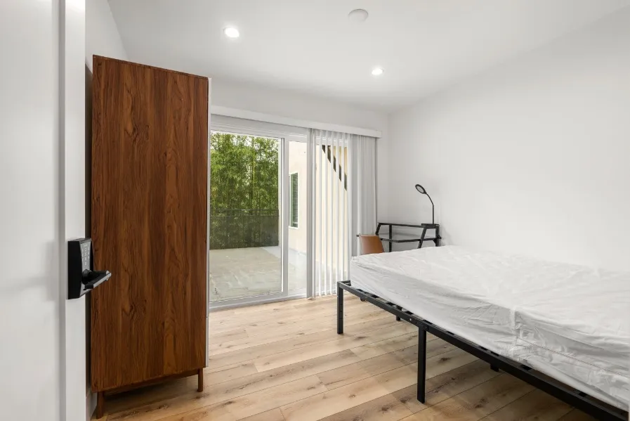 Bright bedroom with wooden floors, featuring a sleek bed frame, a stylish wooden wardrobe, and large sliding glass doors that open to a private balcony with a serene view.