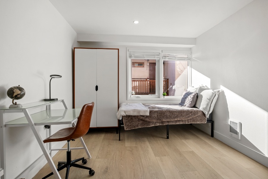 Bright and cozy bedroom featuring a comfortable bed, modern desk with a globe lamp, and natural light streaming through large windows.