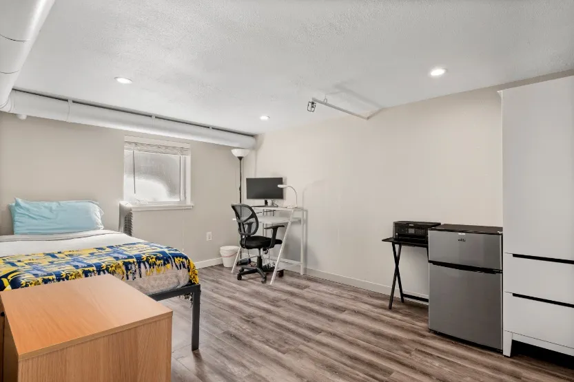 Cozy, modern bedroom with bright natural light, stylish wood flooring, compact workspace, and mini fridge.